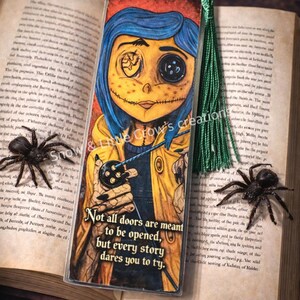 May include: A Coraline-themed bookmark featuring the character with blue hair and a button eye, wearing a yellow coat. The bookmark has a green tassel and the text "Not all doors are meant to be opened, but every story dares you to try."