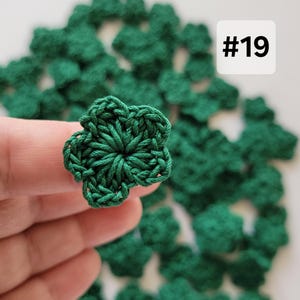 May include: A collection of small, dark green crocheted four-leaf clover shapes. Each clover is crafted from yarn with a textured, looped design. A white square label with the black text #19 is visible in the upper right corner of the image.