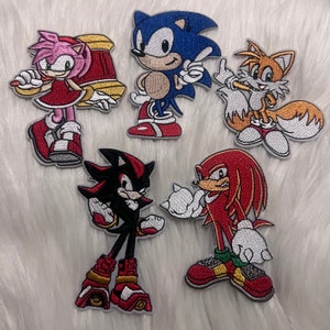 May include: Five embroidered patches featuring characters from the Sonic the Hedgehog video game franchise. The patches are on a white fuzzy background. The characters are Sonic, Tails, Knuckles, Shadow, and Amy.