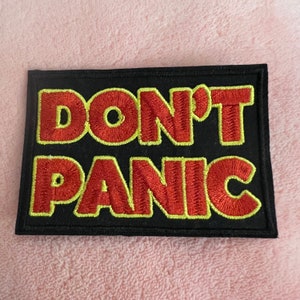 May include: A black fabric patch with yellow stitching around the edges. The patch has the words "DON'T PANIC" embroidered in red.