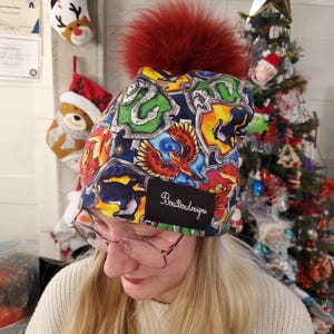 Puede incluir: Un gorro colorido con un gran pompón rojo. El gorro presenta un patrón vibrante con varios elementos gráficos y el texto "BouBoudesigns" en una etiqueta negra. Se ven decoraciones navideñas en el fondo.