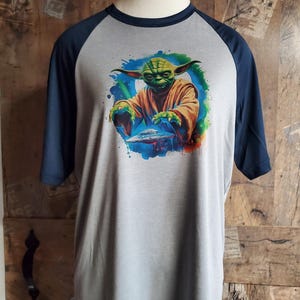 Peut inclure: T-shirt gris à manches raglan avec une illustration colorée de Yoda de Star Wars. Le graphisme représente Yoda dans un style aquarelle avec un vaisseau spatial. Les manches et l'encolure sont bleu marine.