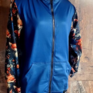 Op de afbeelding: Een blauwe hoodie met ritssluiting en een voorzak. De mouwen en de voering van de capuchon hebben een bloemen- en vossenprint in oranje, zwart en wit. De hoodie wordt op een mannequin gepresenteerd.