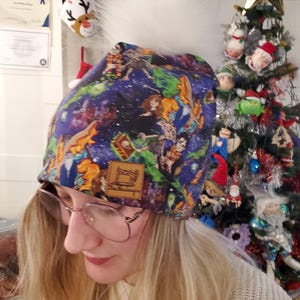 Puede incluir: Un gorro azul con un pompón de piel sintética blanca. El gorro presenta un estampado colorido con varios personajes. Una pequeña etiqueta de cuero marrón con un logotipo de máquina de coser está cosida en la parte delantera.