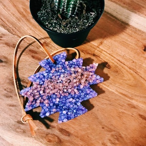 May include: Aromatic air freshener in a starburst shape, filled with blue and pink granules, with a leather loop for hanging. The freshener sits on a wooden surface, next to a potted cactus.