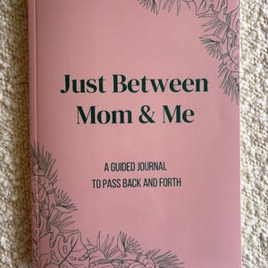 May include: A pink guided journal titled "Just Between Mom & Me." The cover features dark green text and botanical illustrations in the corners. The journal's subtitle reads "A Guided Journal to Pass Back and Forth."