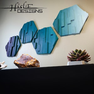 May include: Three teal blue hexagonal wooden wall sculptures with a gradient of color from dark to light. The sculptures are arranged in a staggered pattern on a white wall. The text "Hisle Designs" is visible in the top left corner of the image.