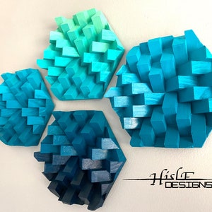 May include: Four hexagonal wall decor pieces made of wood, painted in shades of blue and green. The pieces are arranged in a staggered pattern on a white wall. The text "Histle Designs" is visible in the bottom right corner of the image.