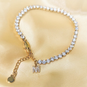 May include: A gold chain bracelet with a single initial charm. The bracelet is made of small, clear cubic zirconia stones.