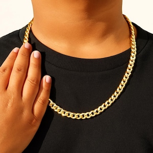 May include: A gold-colored chain necklace with a curb link design. The necklace is worn around the neck and rests on a black shirt. The chain is made of a shiny, reflective metal.