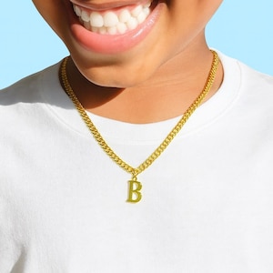 May include: A gold-colored chain necklace with a pendant featuring the letter "B". The necklace is worn over a white t-shirt. The chain has a curb link design.