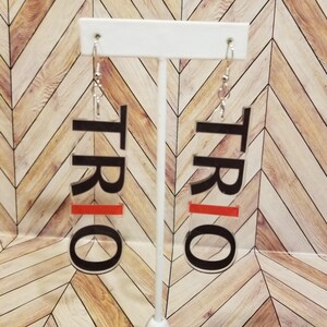 May include: Clear acrylic earrings with the word "TRIO" in black block letters. A horizontal red stripe is below the "TRIO" text. The earrings hang from silver-colored fish hook ear wires.