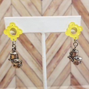 May include: A pair of earrings featuring a yellow flower-shaped stud with a silver teapot charm dangling below. The earrings are displayed on a white earring stand against a wooden background.