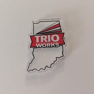 May include: Clear acrylic pin in the shape of the state of Indiana. The pin features the text "TRIO WORKS" in white on a red banner, with a white and red design above. The pin is on a white background.