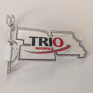 May include: Clear acrylic pin in the shape of a state with the words "TRIO WORKS" in black and red. A silver-colored pin back is attached to the acrylic piece. The pin is on a white surface.