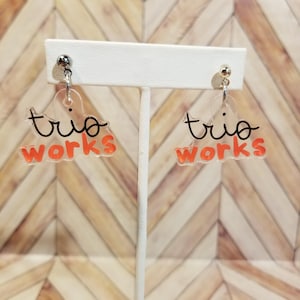 May include: Clear acrylic earrings with the words "trio works" in black script and orange block letters. The earrings are attached to silver-colored posts. The earrings are displayed on a white stand against a wood-grain background.