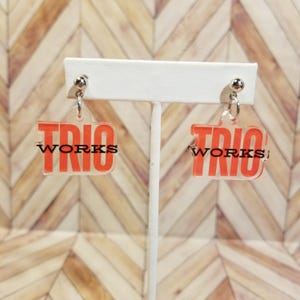 May include: A pair of clear acrylic earrings with a red rectangular design. The earrings have the word "TRIO" in red block letters above the word "WORKS" in black. The earrings are displayed on a white stand against a light brown background.
