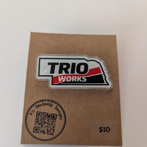 May include: A rectangular pin with the text "TRIO WORKS" in black letters on a white background, with a red and black accent. The pin is attached to a brown card with a QR code and the words "AZ Handmade Designs".