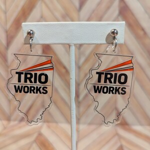 May include: Clear acrylic earrings shaped like the state of Illinois. The earrings feature the text "TRIO WORKS" in black, with orange and black angled lines at the top. The earrings hang from silver-colored ball studs.