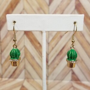 May include: A pair of gold dangle earrings with green cactus charms. The cactus charms have a white outline and are set in a gold pot with clear rhinestones.