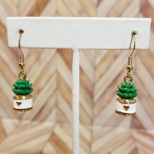 May include: A pair of gold dangle earrings with a green potted plant charm. The plant is in a white pot with a gold heart on the front.