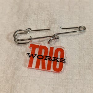 May include: A silver-colored safety pin with a clear plastic tag that reads "TRIO WORKS" in red block letters. The pin is open and resting on a white, textured surface.