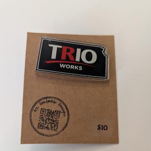 May include: A rectangular pin featuring the text "TRIO WORKS" in white and red on a black background, shaped like the state of Kansas. The pin is attached to a brown card with a circular stamp and the text "AZ Handmade Designs".