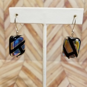 May include: A pair of silver dangle earrings with iridescent glass rectangular beads. The beads have a black geometric pattern.