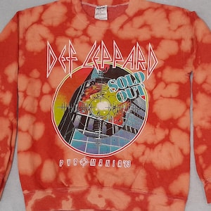 Könnte beinhalten: Rot-oranges Sweatshirt mit Batikmuster und der Band Def Leppard. Auf der Vorderseite steht der Bandname in weißen, eckigen Buchstaben über einer runden Grafik mit den Worten "SOLD OUT" und "PYROMANIA '83".