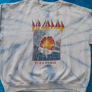 Könnte beinhalten: Hellgraues Sweatshirt mit Batikmuster, mit dem Def Leppard-Logo und dem Wort "PYROMANIA" in Rot. Die Grafik zeigt ein rotes und gelbes Zielscheibendesign. Das Sweatshirt hat lange Ärmel und einen Rundhalsausschnitt.