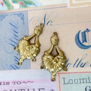 May include: Two gold-toned metal embellishments with a decorative floral design and three loops. The embellishments are on a white background with blue text.