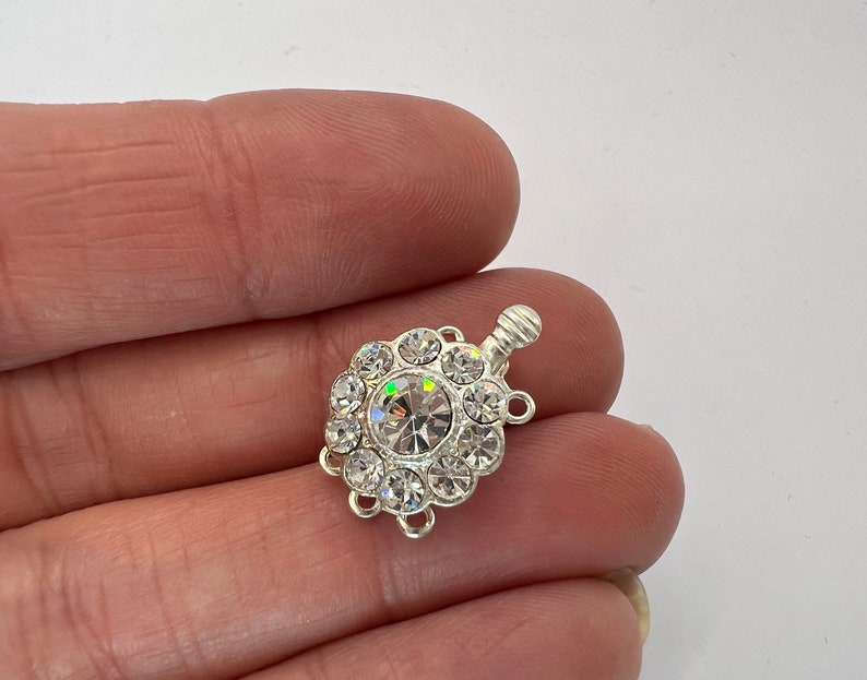May include: Silver-toned clasp with a round crystal center surrounded by smaller crystals. The clasp has three loops for attaching to jewelry.