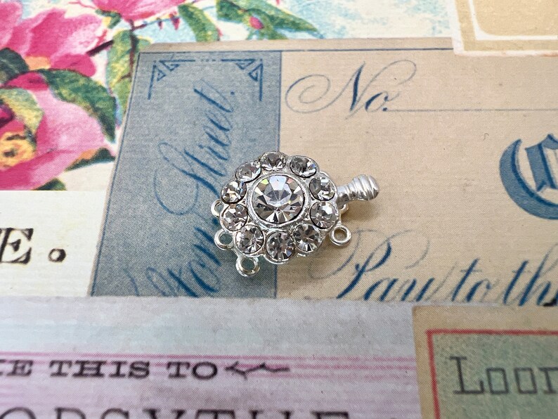 May include: Silver tone clasp with a clear rhinestone center surrounded by smaller rhinestones. The clasp has three loops for attaching to a necklace or bracelet.