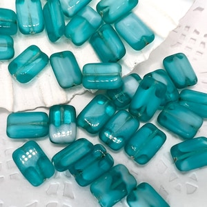 Könnte beinhalten: Eine Sammlung von tealblauen Glasperlen mit einem matten weißen Finish. Die Perlen sind rechteckig und haben eine glatte, polierte Oberfläche.