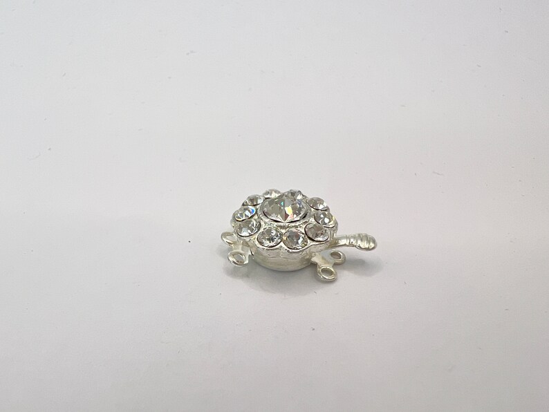 May include: Silver-toned clasp with a round design featuring clear rhinestones. The clasp has three loops for attaching jewelry.