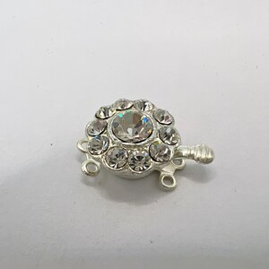 May include: Silver-toned clasp with a round crystal center surrounded by smaller crystals. The clasp has three loops for attaching jewelry.
