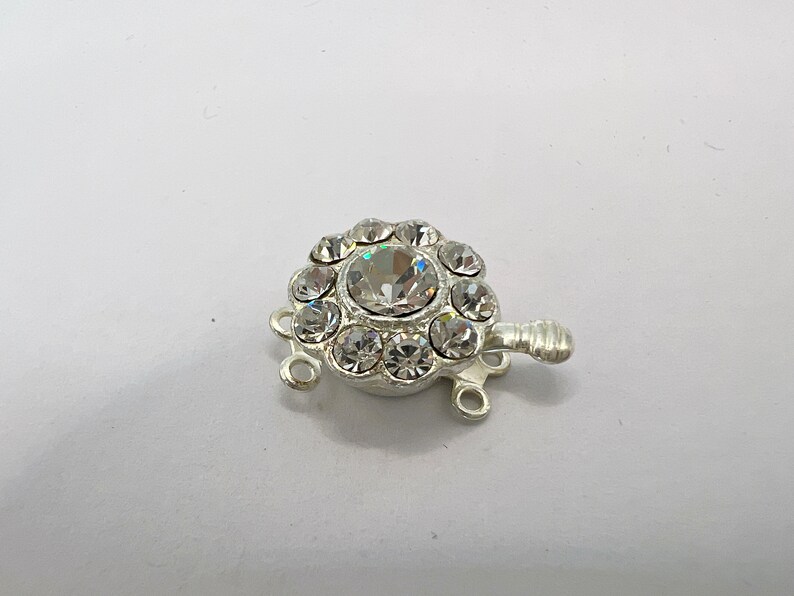 May include: Silver-toned clasp with a round crystal center surrounded by smaller crystals. The clasp has three loops for attaching jewelry.