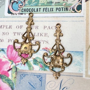 May include: Two gold-toned metal charms with a floral design. Each charm features a stylized flower with a center and petals, surrounded by intricate scrollwork. The charms have a loop at the top for attaching to a necklace or bracelet.