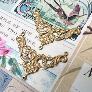 Puede incluir: Dos piezas de esquina decorativas de metal dorado con intrincados diseños de volutas. Las piezas están sobre una superficie estampada con texto que dice "SAMPLE OF OUR PA NO TWO 50 FOR 10 JKE, Colibri paon NOB".