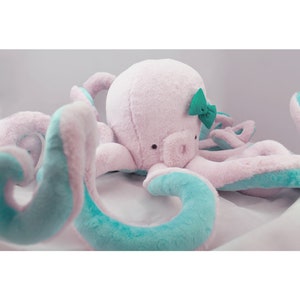 Peut inclure: Une pieuvre en peluche rose avec des tentacules turquoise et un nœud vert. La pieuvre a des yeux noirs et une expression mignonne et amicale.