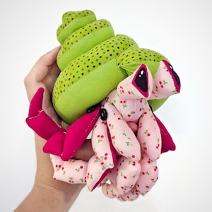Puede incluir: Un juguete de peluche de cangrejo ermitaño con una concha verde con lunares blancos y un cuerpo rosa con estampado de cerezas. El cangrejo está sosteniendo sus garras juntas.