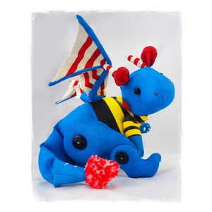May include: A handmade blue plush dragon with red and white striped wings and accents. The dragon has a yellow and black striped belly, red ears, and button eyes. A red pom-pom is attached to the dragon's tail.