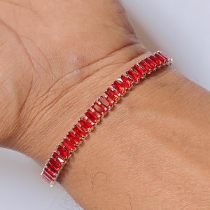 May include: A silver bracelet with rectangular, vibrant red gemstones. The bracelet is worn on a wrist, showcasing the jewelry's design and color. The gemstones are closely set, creating a continuous line of red.