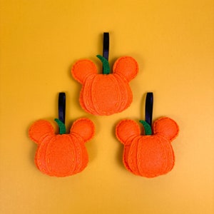 May include: Three orange felt pumpkins with green stems and black string hangers. The pumpkins are shaped like Mickey Mouse heads. The pumpkins are on a yellow background.