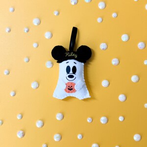 Op de afbeelding: Een wit vilten spookvormig Halloween-ornament met zwarte Mickey Mouse-oren en een zwart lint. De geest houdt een oranje pompoen vast. De naam "Riley" is geborduurd op de zwarte oren. De achtergrond is geel met witte, iriserende, cirkelvormige decoraties.