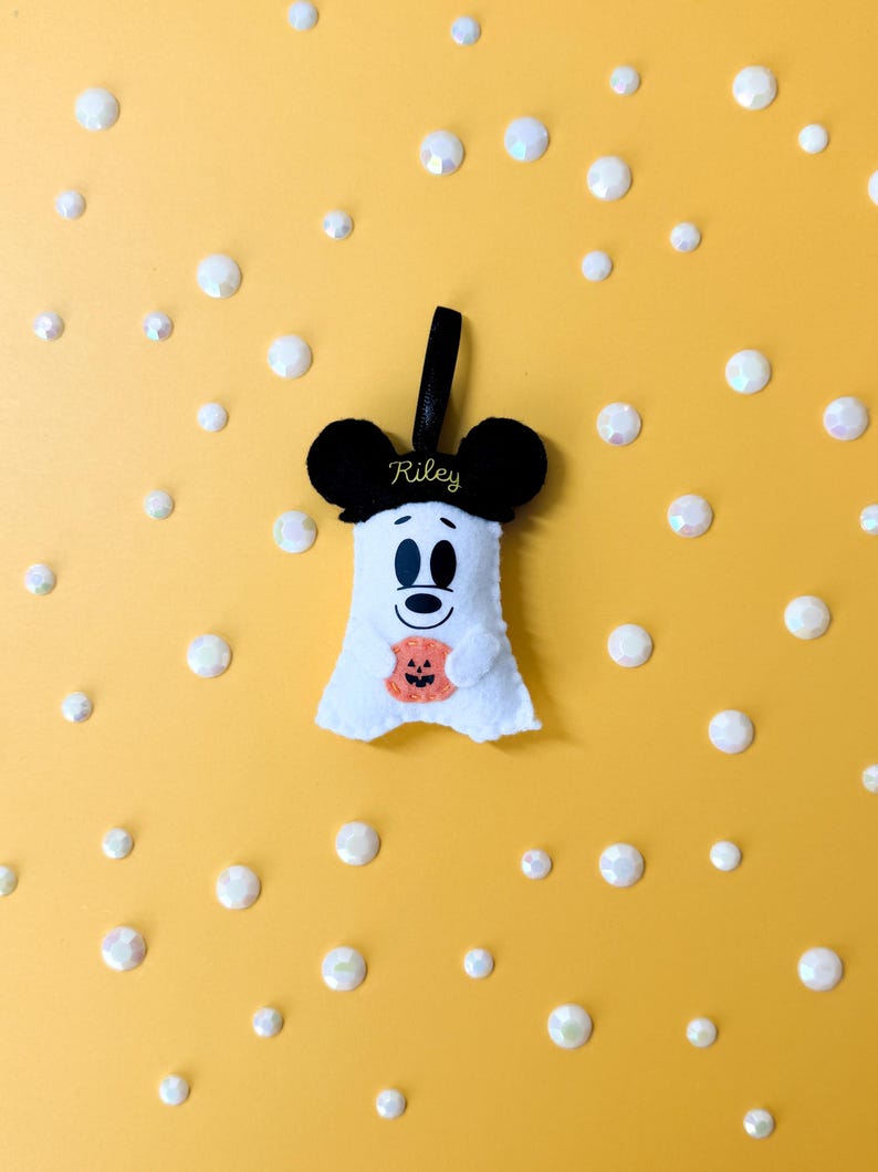 Op de afbeelding: Een wit vilten spookvormig Halloween-ornament met zwarte Mickey Mouse-oren en een zwart lint. De geest houdt een oranje pompoen vast. De naam "Riley" is geborduurd op de zwarte oren. De achtergrond is geel met witte, iriserende, cirkelvormige decoraties.