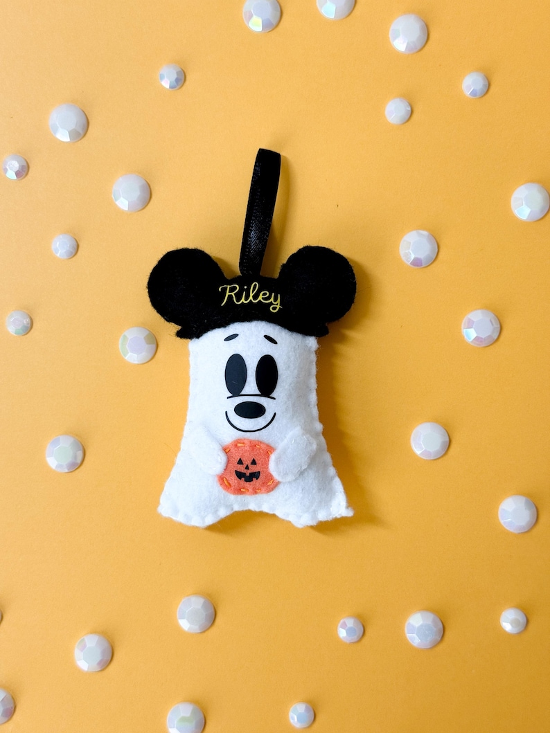 Op de afbeelding: Een wit vilten spookvormig ornament met zwarte muizenoren en een zwart lint om op te hangen. De geest houdt een oranje pompoen vast en heeft de naam "Riley" geborduurd op de zwarte hoed. De achtergrond is geel met verspreide witte iriserende edelstenen.