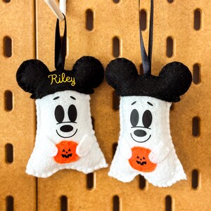 Op de afbeelding: Twee witte vilten spookornamenten met zwarte muizenoren en zwarte linten om op te hangen. Elk spook houdt een oranje pompoen vast en heeft een lachend gezicht. E&eacute;n ornament heeft de naam "Riley" in geel geborduurd.
