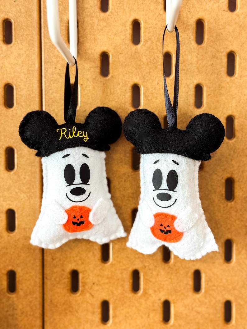 Op de afbeelding: Twee witte vilten spookornamenten met zwarte muizenoren en zwarte linten om op te hangen. Elk spook houdt een oranje pompoen vast en heeft een lachend gezicht. E&eacute;n ornament heeft de naam "Riley" in geel geborduurd.