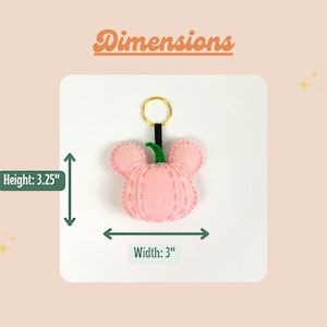 May include: A pink felt pumpkin keychain with a green stem and gold key ring. The keychain measures 3.25 inches in height and 3 inches in width.